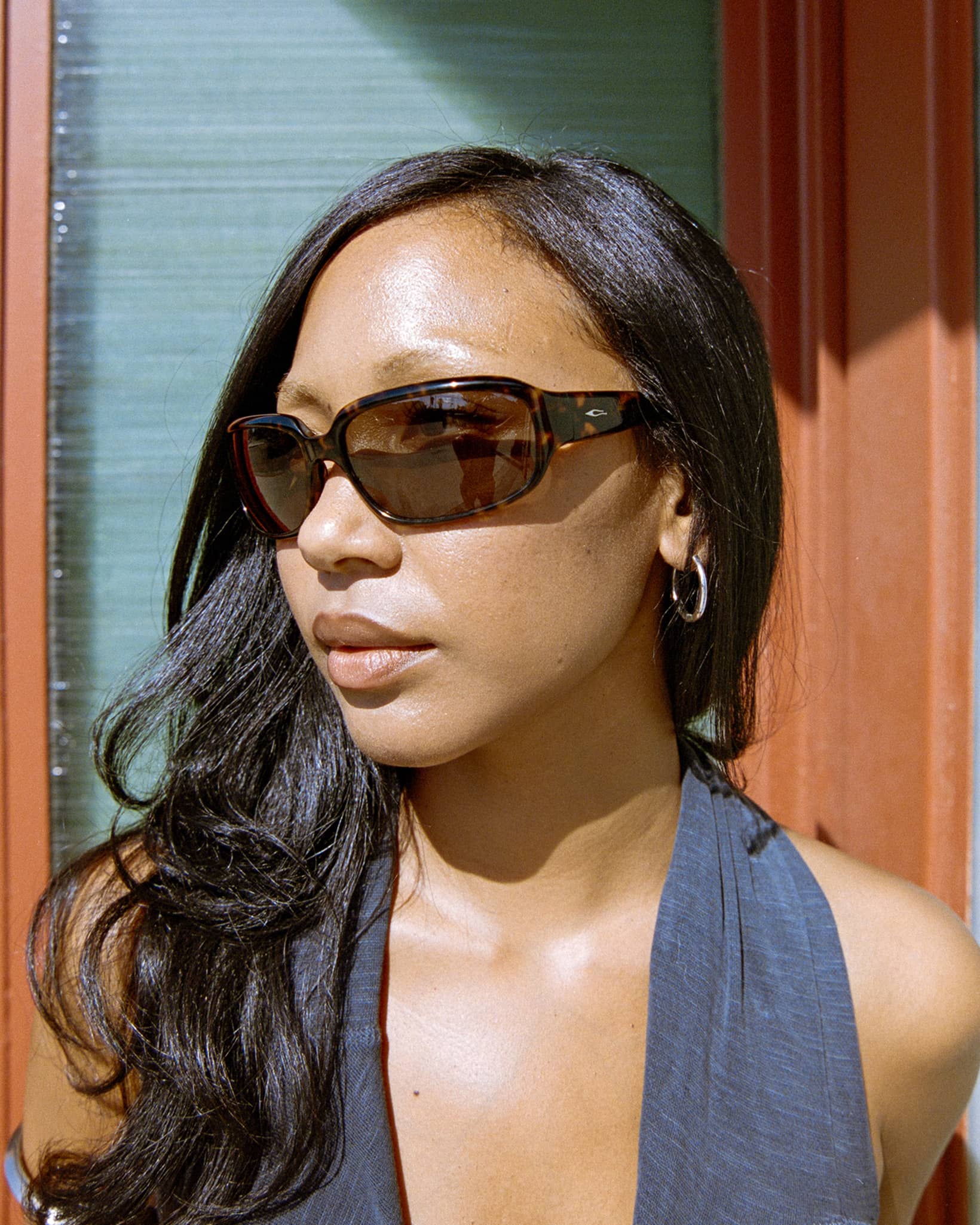 A woman with long dark hair wearing dark brown cat-eye sunglasses and a gray halter top, photographed in warm sunlight against a wooden and glass background.-The Stone Echo - Dark Tortoise Bio-3