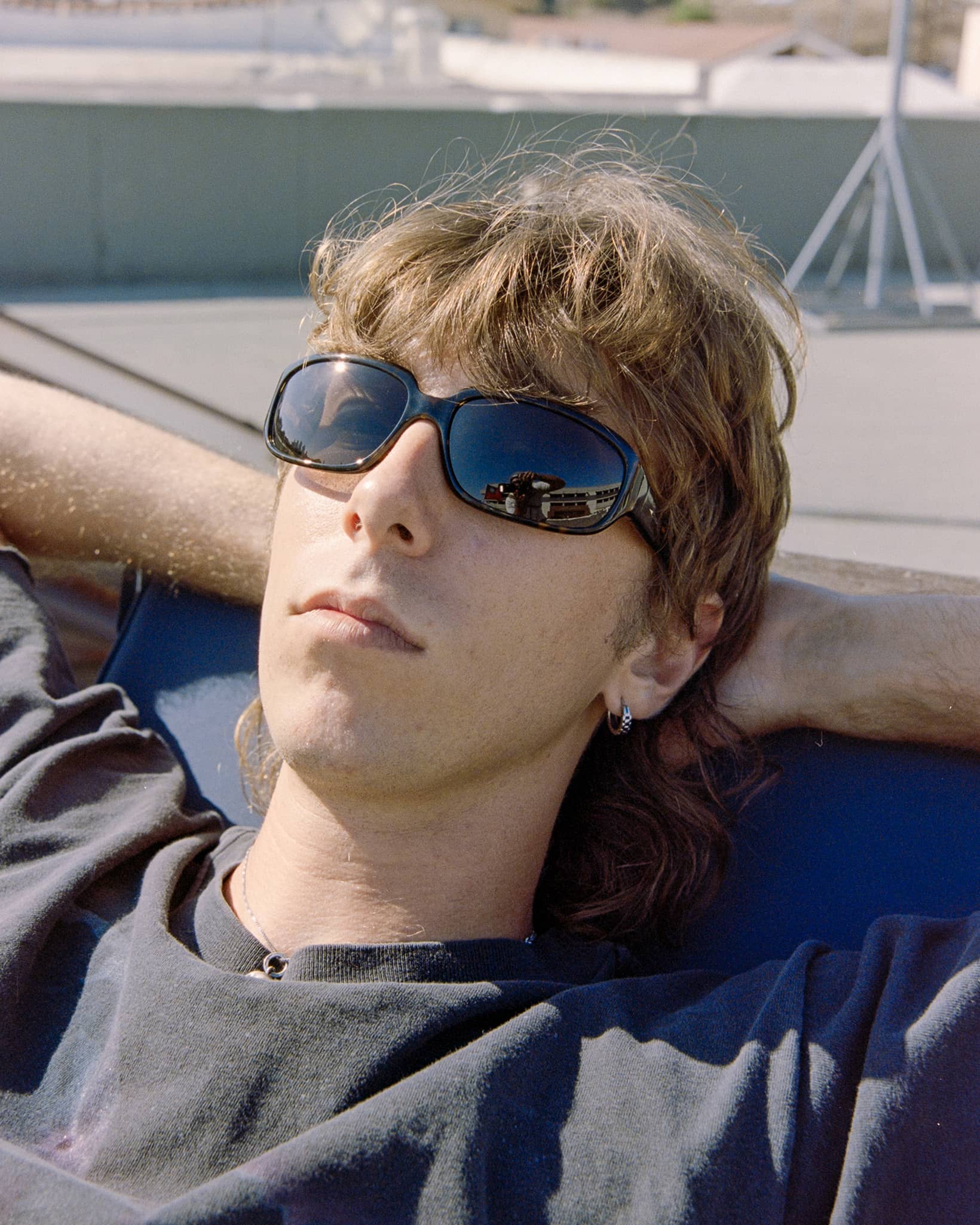 A close-up lifestyle shot of a young man with light brown hair wearing black sunglasses and a dark gray t-shirt, reclining outdoors on a blue lounge chair.-The Stone Echo - Dark Tortoise Bio-4