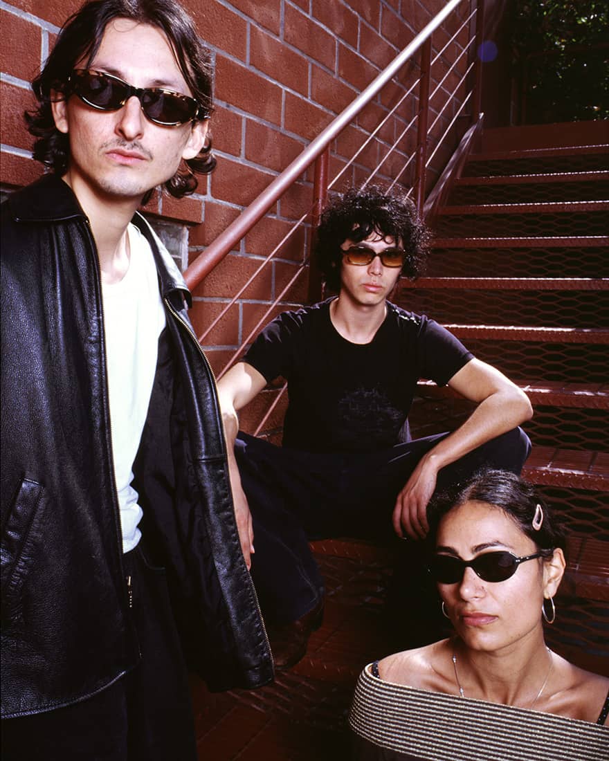 Three people in sunglasses posing on a staircase against a red brick wall